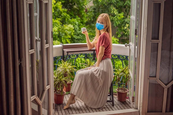 Una donna dai capelli rossi che indossa una maschera medica durante il COVID-19 coronavirus si trova su un balcone in stile heritage che si gode il suo caffè mattutino. Una donna in un hotel in Europa o in Asia come il turismo si riprende da un — Foto Stock