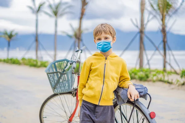 Активний блондин хлопець у медичній масці під час коронавірусу COVID-19 їде на велосипеді в парку біля моря. Дитина Тоддлер мріє і веселиться в теплий літній день. ігри на відкритому повітрі для дітей — стокове фото