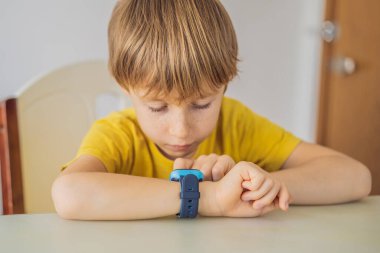 Küçük çocuk masada oturuyor ve akıllı görünüyor. Bebek güvenliği için akıllıca bir saat. Çocuk okul dersleri alıyor, müzik dinliyor, arkadaşlarını arıyor.