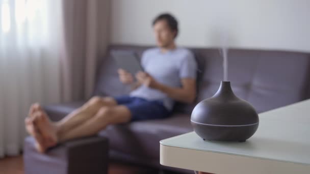 The man in the background is sitting on a couch and a working aromatherapy diffuser is on a table in a foreground — Stock Video