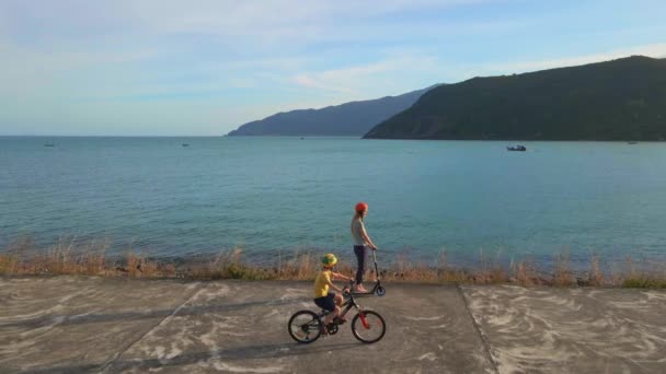 Повільний повітряний постріл. Жінка та її син катаються на скутері та велосипеді біля моря під час заходу сонця — стокове відео