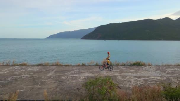 Disparo aéreo en cámara lenta. Mujer y su hijo montan patada scooter y bicicleta junto al mar durante la puesta del sol — Vídeos de Stock