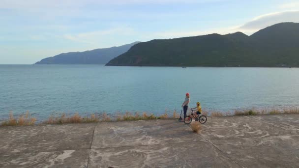 Zeitlupenaufnahme aus der Luft. Frau und ihr Sohn fahren Tretroller und Fahrrad am Meer bei Sonnenuntergang — Stockvideo