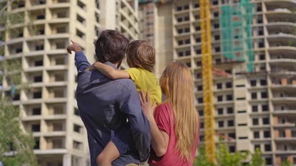 Un padre, una madre y un hijo de familia miran un edificio alto que está en construcción. Están discutiendo dónde deberían comprar un apartamento. — Vídeos de Stock
