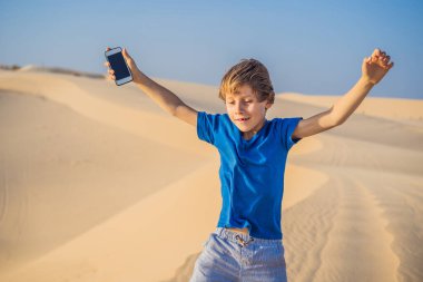 Çölde elinde akıllı bir telefonla dans eden sevimli, komik çocuk.