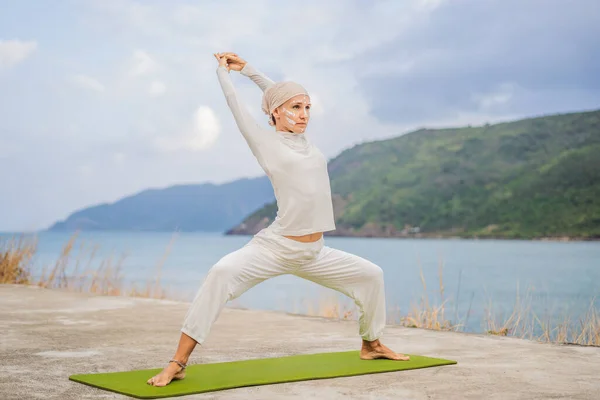 Kundalini yoga woman in white clothes and turban practices yoga kundalini on the background of the sea, mountains and sunset. Fighting face painting of the Indians shows her inner world. Visual