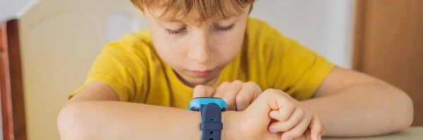 BANNER, LONG FORMAT Liten pojke sitter vid bordet och ser smart klocka. Smart klocka för babysäkerhet. Barnet gör skollektioner, lyssnar på musik, ringer vänner — Stockfoto