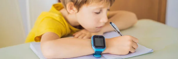 BANNER, LONG FORMAT Little boy sitting at the table and looking smart watch. Smart watch for baby safety. The child makes school lessons, listening to music, calling friends — Stock Photo, Image
