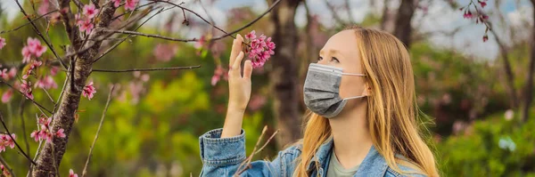 Mujer feliz en sakura en máscara protectora y oliendo flores de sakura florecientes después de la cuarentena del coronavirus BANNER, FORMATO LARGO — Foto de Stock