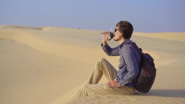 A man sitting on a dune in a desert drinks water from a reusable bottle — Stock Video