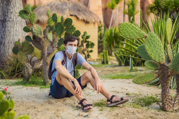 Beautiful stylish young man in desert among the cacti, wearing a medical mask during COVID-19 coronavirus, traveling in Africa on safari, exploring nature, sunny summer, traveler on vacation — Stock Photo, Image