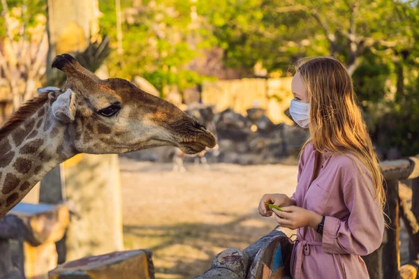 Happy young woman watching and feeding giraffe in zoo wearing a medical mask during COVID-19 coronavirus. Happy young woman having fun with animals safari park on warm summer day — Stock Photo, Image