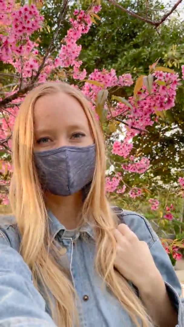 Vertikales Video. Voman mit Gesichtsmaske macht Selfie-Video, das unter einem blühenden Sakura-Baum steht. Reisen Sie nach Japan in Zeiten von Pandemien. Schuss auf ein Handy — Stockvideo