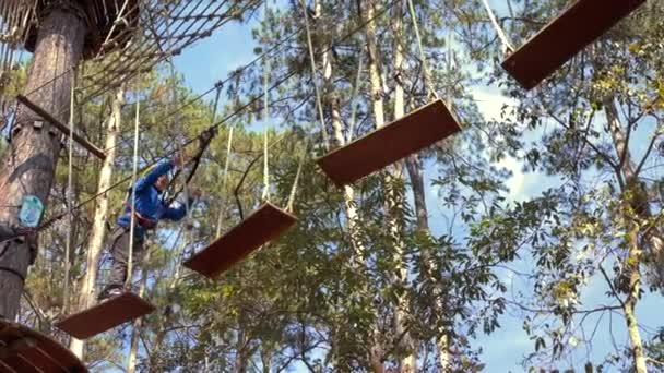 Filmagem em câmera lenta de um menino em um capacete amarelo que está se divertindo em um parque de aventura de escalada de corda. Tiro em um telefone — Vídeo de Stock