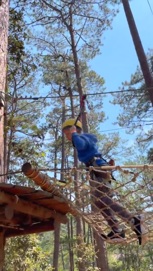 Vídeo vertical. Slowmotion tiro de um menino em um capacete amarelo que está se divertindo em um parque de aventura escalada corda — Vídeo de Stock