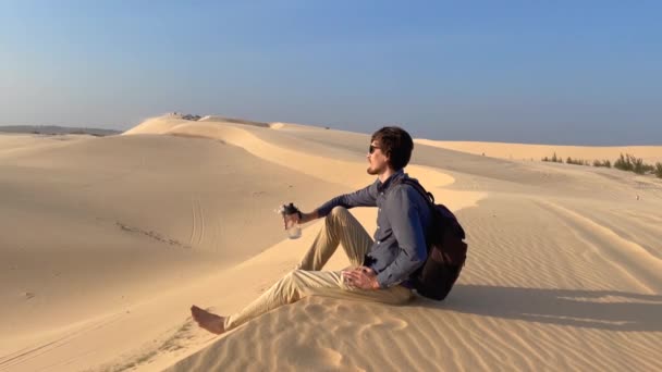 Fotografía en cámara lenta de un hombre que bebe agua de una botella reutilizable sentado en una duna en un desierto — Vídeos de Stock