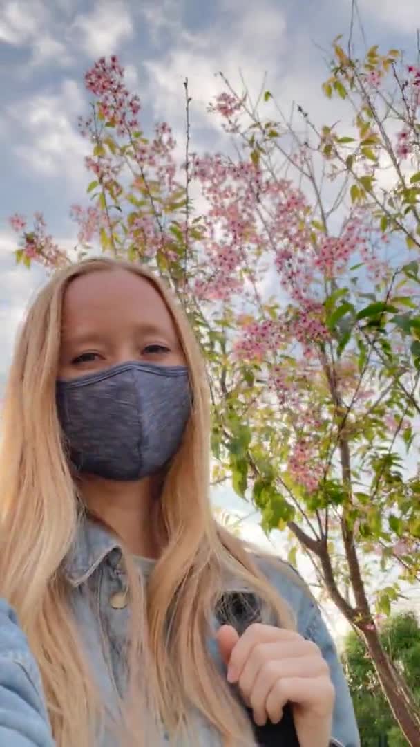 Vertical video. Voman wearing a facemask makes selfie video standing under blooming sakura tree. Travel to Japan in a times of pandemics. Shot on a phone — Stock Video