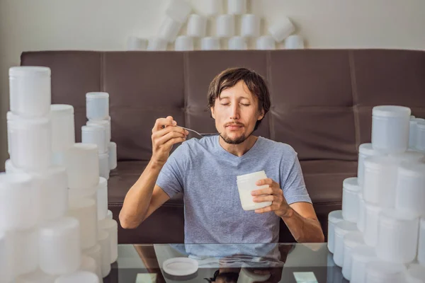 A férfi joghurtot eszik, és sok üveg joghurttal van körülvéve, amit egy év alatt használ. Hatalmas mennyiségű műanyag ételes üveggel. Túlzott műanyagfogyasztás. Csökkentés, újrafelhasználás, újrahasznosítás — Stock Fotó
