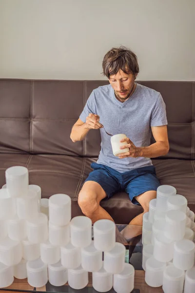 The man eats yogurt and is surrounded by many jars of yoghurt that he uses in a year. A huge amount of plastic food jars. Excessive consumption of plastic. Reduce, reuse, recycle — Stock Photo, Image