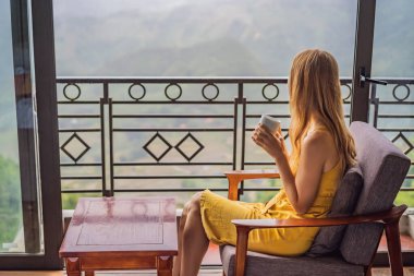 Balkonda sandalyeye oturmuş, dağlara ve yeşil doğaya bakan genç bir kadın kahve fincanı tutuyor. Sapa 'da siste, Kuzeybatı Vietnam' da bir kadın. Vietnam seyahat konsepti. UNESCO Mirası