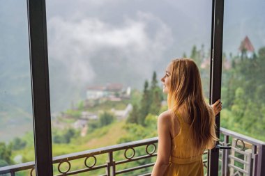 A young woman holding coffee cup while sittinhg on chair on balcony , looking at mountains and green nature. Woman in Sapa in the fog, Northwest Vietnam. Vietnam travel concept. UNESCO heritage clipart