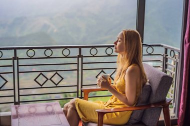 Balkonda sandalyeye oturmuş, dağlara ve yeşil doğaya bakan genç bir kadın kahve fincanı tutuyor. Sapa 'da siste, Kuzeybatı Vietnam' da bir kadın. Vietnam seyahat konsepti. UNESCO Mirası
