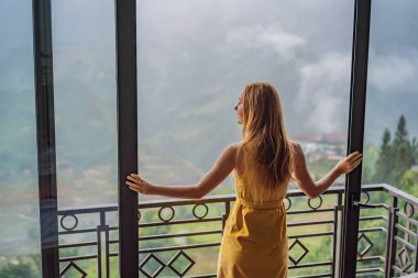 Balkonda sandalyeye oturmuş, dağlara ve yeşil doğaya bakan genç bir kadın kahve fincanı tutuyor. Sapa 'da siste, Kuzeybatı Vietnam' da bir kadın. Vietnam seyahat konsepti. UNESCO Mirası
