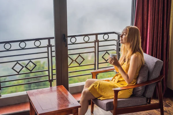 Una giovane donna che tiene in mano una tazza di caffè mentre siede sulla sedia sul balcone, guardando le montagne e la natura verde. Donna a Sapa nella nebbia, Vietnam nord-occidentale. Vietnam concetto di viaggio. Patrimonio UNESCO — Foto Stock