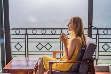 Balkonda sandalyeye oturmuş, dağlara ve yeşil doğaya bakan genç bir kadın kahve fincanı tutuyor. Sapa 'da siste, Kuzeybatı Vietnam' da bir kadın. Vietnam seyahat konsepti. UNESCO Mirası