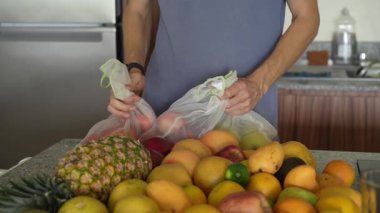 Genç bir adam sağlıklı ve renkli bir düzenleme hazırlamak için mutfaktaki bir masaya taze meyve koyar. Parlak ve davetkar bir yerde ev yapımı, sağIık ve düşünceli bir yaşam..