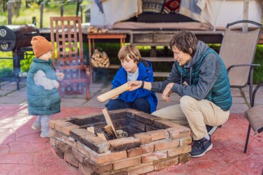 Baba ve iki oğlu bir kamp alanı önünde kamp ateşi yakıyor aile bağlarının, açık hava maceralarının ve doğa keşiflerinin keyfini çıkarıyorlar..