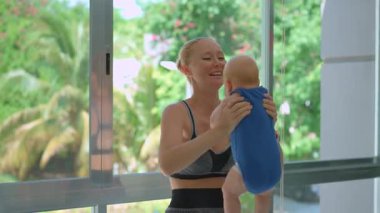 Genç anne, arka planda tropik manzarası olan büyük bir pencerenin yanında 3 aylık oğluyla egzersiz yapıyor..