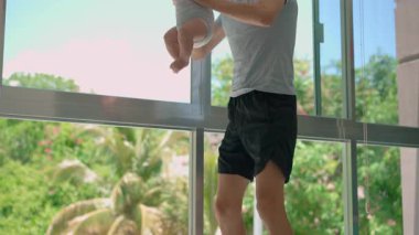 Genç baba, arka planda tropik manzarası olan büyük bir pencerenin yanında 3 aylık oğluyla egzersiz yapıyor..