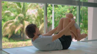 Genç baba, arka planda tropik manzarası olan büyük bir pencerenin yanında 3 aylık oğluyla egzersiz yapıyor..