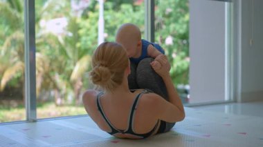 Genç anne, arka planda tropik manzarası olan büyük bir pencerenin yanında 3 aylık oğluyla egzersiz yapıyor..