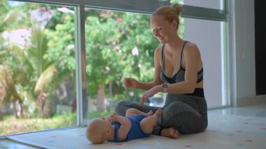 Genç anne, arka planda tropik manzarası olan büyük bir pencerenin yanında 3 aylık oğluyla egzersiz yapıyor..