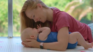 Annem 3 aylık bebeğini tropikal arka planı olan büyük bir pencerenin yanında öpüyor. Şefkat bağı ve erken annelik kavramı.