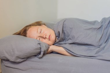 Kadın, yumuşak gri bir yatak ile yatakta huzur içinde uyuyor, sakin ve rahat bir sabah atmosferinin tadını çıkarıyor. Dinlenme, rahatlama ve refah kavramı, sağlıklı uykuyu, huzuru ve konforu temsil eder.