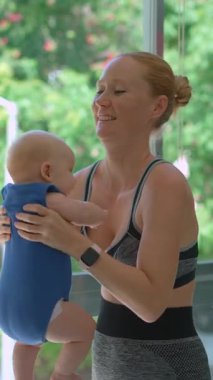 Genç anne, arka planda tropik manzarası olan büyük bir pencerenin yanında 3 aylık oğluyla egzersiz yapıyor..