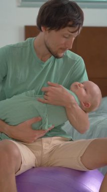 Bir baba kundaklanmış yeni doğmuş bebeğini bir fitbol topunun üzerinde otururken nazikçe sallayarak uyutur. Bebek sakin ve rahatlamış. Huzurlu bir ebeveyn, bağ kurma ve yatma zamanı bakımı sahnesi.