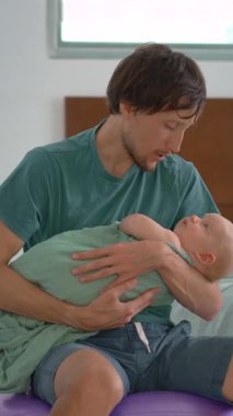 Bir baba kundaklanmış yeni doğmuş bebeğini bir fitbol topunun üzerinde otururken nazikçe sallayarak uyutur. Bebek sakin ve rahatlamış. Huzurlu bir ebeveyn, bağ kurma ve yatma zamanı bakımı sahnesi.