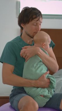 Bir baba kundaklanmış yeni doğmuş bebeğini bir fitbol topunun üzerinde otururken nazikçe sallayarak uyutur. Bebek sakin ve rahatlamış. Huzurlu bir ebeveyn, bağ kurma ve yatma zamanı bakımı sahnesi.