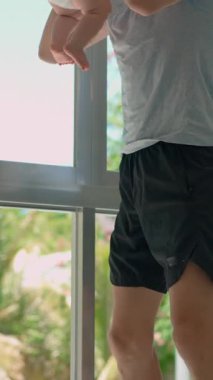 Genç baba, arka planda tropik manzarası olan büyük bir pencerenin yanında 3 aylık oğluyla egzersiz yapıyor..