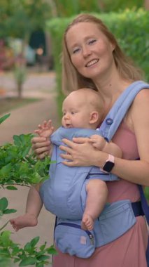 Genç anne, güneşli bir günde bebeğiyle parka gidip ergonomik bir taşıyıcıyla kaliteli zaman ve doğanın tadını çıkarıyor..