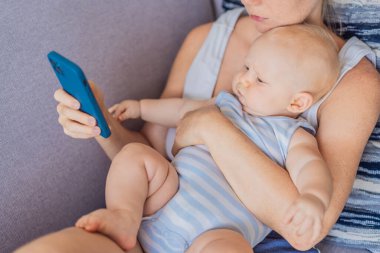 Bebek akıllı telefonuna bakarken annesinin kucağında oturuyor, duygusal olarak kopuk. Perdenin bebekler ve karşılanmamış duygusal ihtiyaçlar üzerindeki etkisini gösteren sembolik bir sahne. Dijital ebeveynlik ve