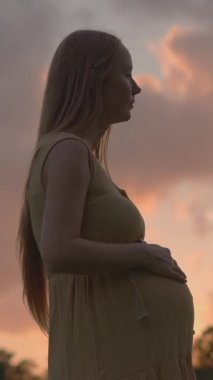 Hamile bir kadın, canlı turuncu ve mor günbatımı gökyüzünün güzelliğiyle çevrili bir tarlada tek başına duruyor. Huzurlu, duygusal annelik sahnesi doğayı, dinginliği ve yeni bir beklentiyi simgeliyor.