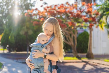 Bir anne, ergonomik bir bebek taşıyıcısı ile bebeğini vücuduna yakın bir şekilde dışarı çıkar. Bebek giyimi, aktif ebeveynlik, rahatlık, bağlılık, hareketlilik ve modern aile yaşam tarzı kavramı
