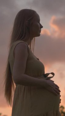 Hamile bir kadın, canlı turuncu ve mor günbatımı gökyüzünün güzelliğiyle çevrili bir tarlada tek başına duruyor. Huzurlu, duygusal annelik sahnesi doğayı, dinginliği ve yeni bir beklentiyi simgeliyor.