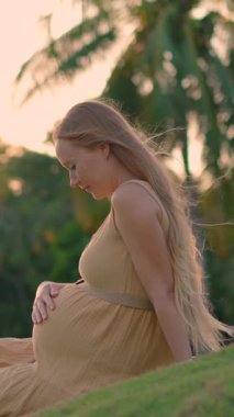 Hamile kadın gün batımında arkasında tropikal ağaçlarla çimenlerde oturur. Doğadaki huzurlu anın tadını çıkarır ve hamileliğini kucaklar..