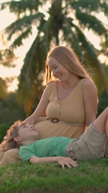 Hamile bir kadın ve büyük oğlu, renkli bir günbatımı gökyüzünün altında palmiye ağaçlarıyla çevrili bir tepede birlikte oturuyorlar. Serene aile bağı, duygusal bağ ve tropikal annelik yaşam tarzı